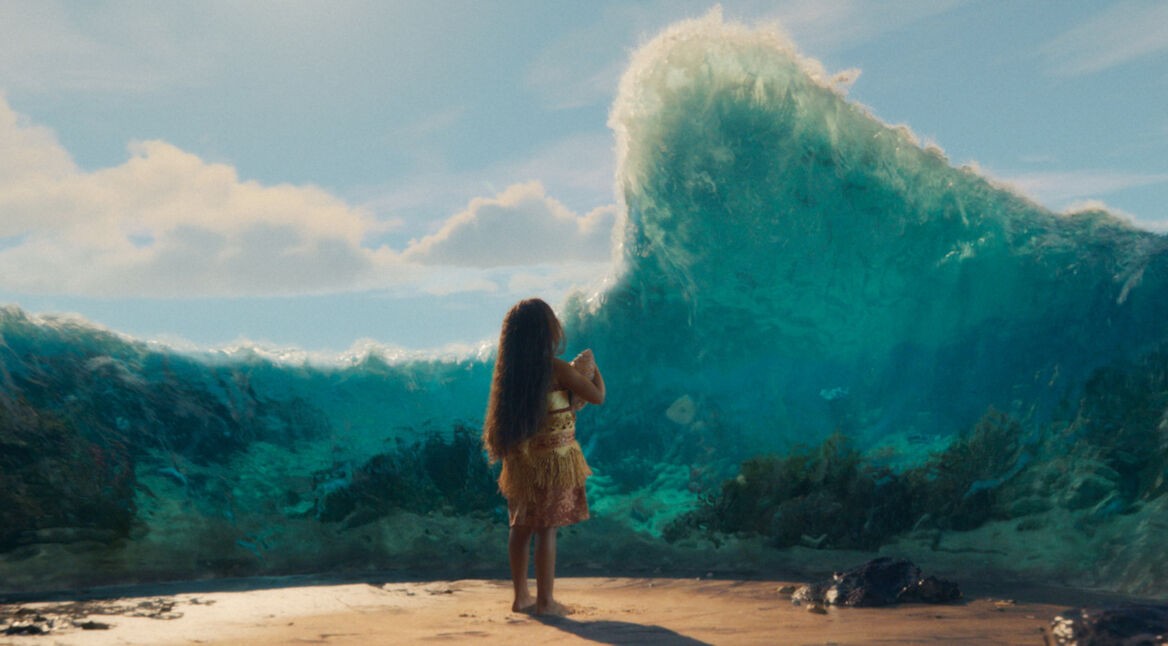 In dieser Szene aus der Vaiana-Realverfilmung steht ein kleines Mädchen mit langen, offenen Haaren barfuß am Strand und blickt auf eine gewaltige Wasserwand, die sich vor ihr auftürmt.