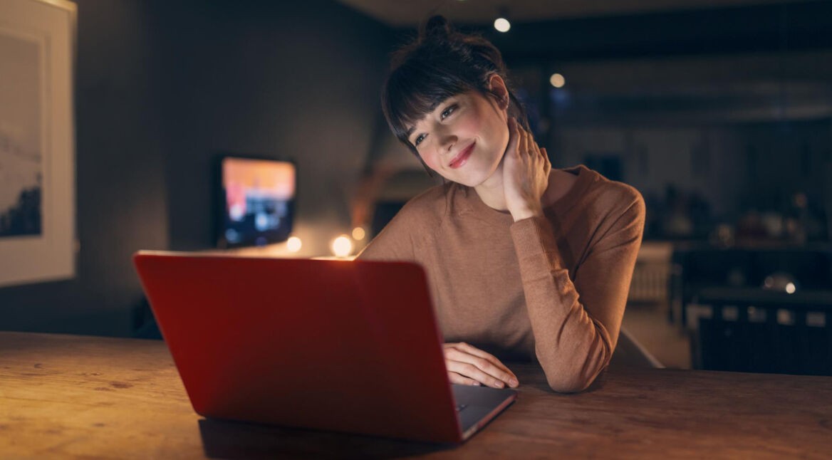Auf dem Bild zum Vodafone Speedtest sitzt eine junge Frau entspannt an einem Holztisch vor einem roten Laptop in einer gemütlich beleuchteten Wohnung. Sie lächelt leicht und blickt konzentriert auf den Bildschirm. Die warme Lichtstimmung und der moderne Wohnraum im Hintergrund vermitteln eine ruhige und angenehme Atmosphäre, passend zu schnellem und komfortablem Internet zuhause.