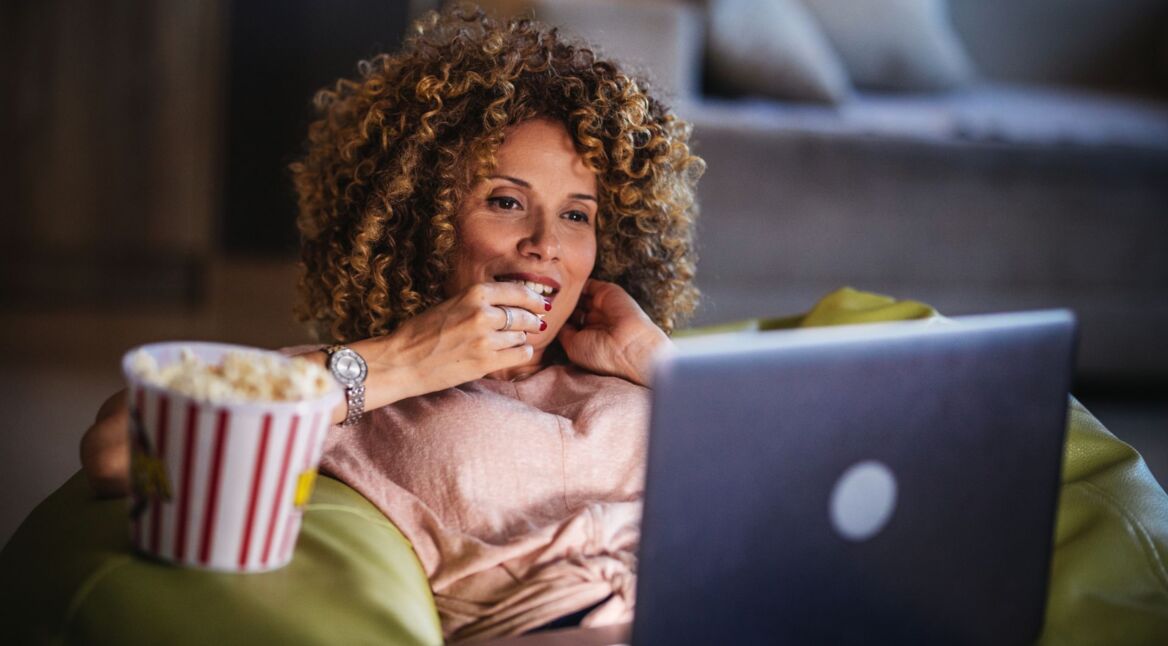 Schwarze Frau sitzt auf der Couch im Wohnzimmer und streamt mit ihrem MacBook.