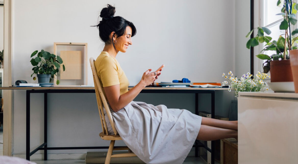 Seitenansicht einer jungen Frau, die am Schreibtisch sitzt und auf ihr Smartphone schaut.
