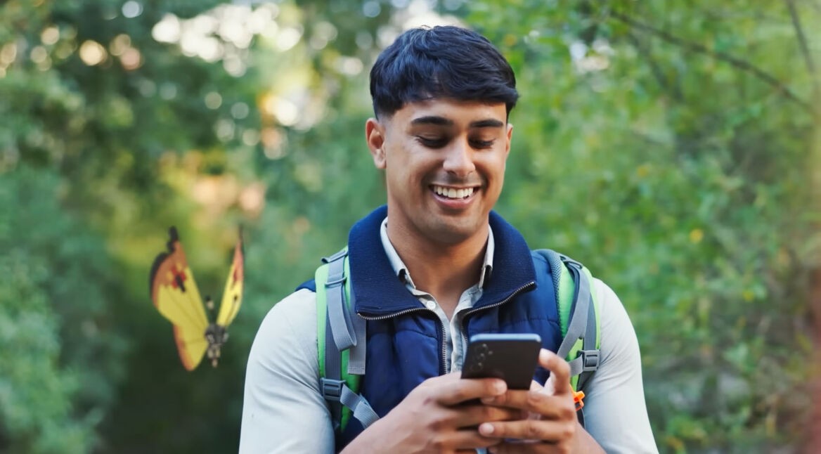 Ein Mann steht im Freien und hat sein Handy in der Hand, während ein Vivilon mit Kontinentalmuster hinter ihm fliegt