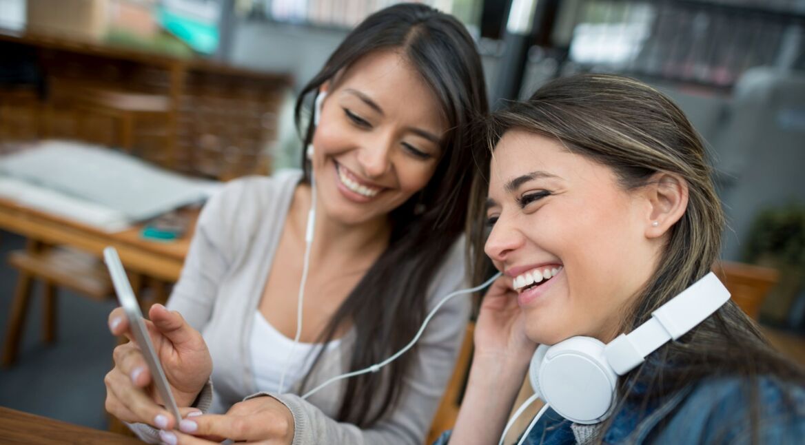 Zwei Frauen teilen sich Kopfhörer und hören gemeinsam Musik mit dem Smartphone.