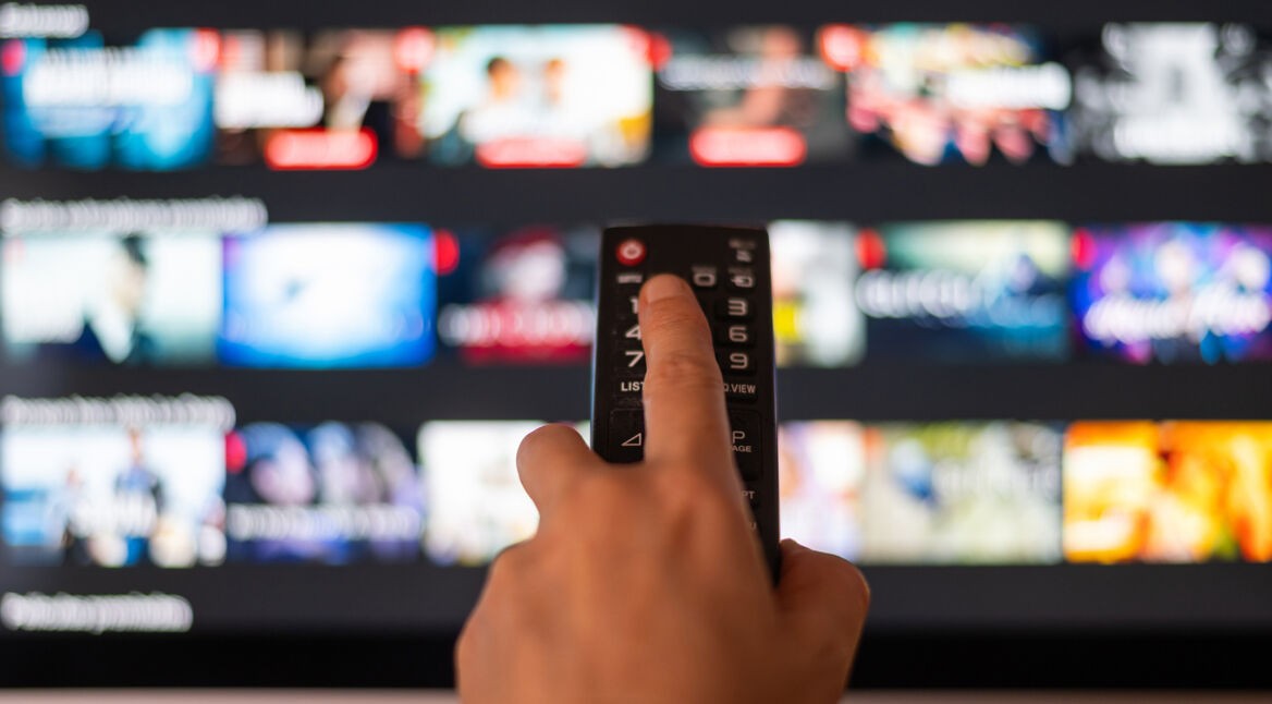 Auf dem Bild in dem Artikel zu den Kosten von Apple TV+ im Jahr 2025 ist eine Hand zu sehen, die eine Fernbedienung hält und auf einen Fernseher zielt. Auf dem Bildschirm sind verschwommene Miniaturansichten von Filmen und Serien in einer Streaming-Oberfläche sichtbar. Die Szene vermittelt eine moderne, digitale Atmosphäre.