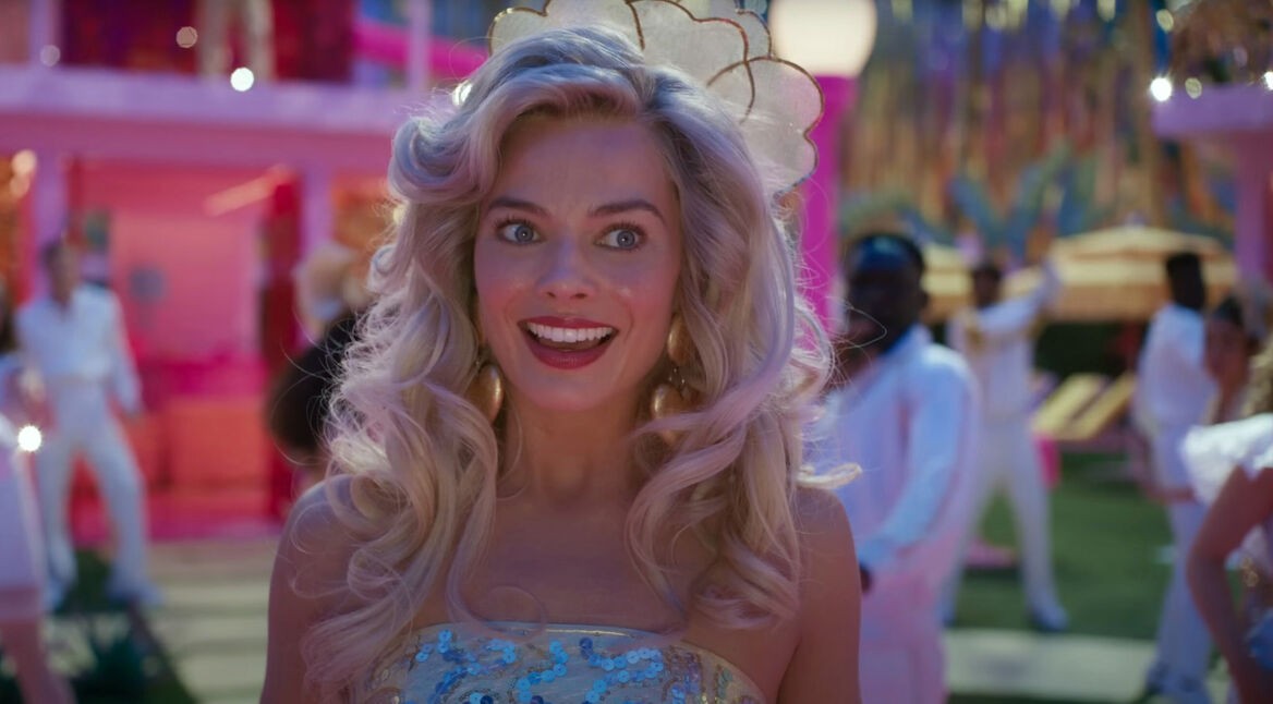 Auf dem Bild zu Barbie 2 ist Barbie, gespielt von Margot Robbie, in einer lebhaften Szene aus dem ersten Barbie-Film zu sehen. Sie trägt ein glitzerndes, mit Pailletten besetztes Kleid und hat voluminöses blondes Haar. Ihr Gesichtsausdruck ist fröhlich und begeistert. Der Hintergrund ist bunt und belebt, mit tanzenden Personen in weißen Outfits und einer festlichen Atmosphäre.