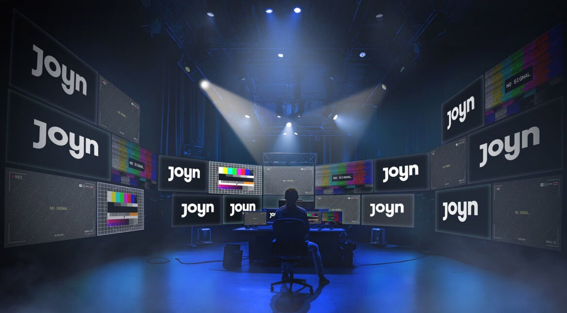 Auf dem Bild zu "Big Brother 2025" sieht man eine futuristische Kontrollzentrale mit zahlreichen Bildschirmen, auf denen teils das Logo von Joyn und teils Testbilder mit der Aufschrift "No Signal" zu sehen sind. In der Mitte sitzt eine Person im Schatten vor einem Pult, das von blauen Scheinwerfern beleuchtet wird. Die Szene wirkt geheimnisvoll und technologisch.