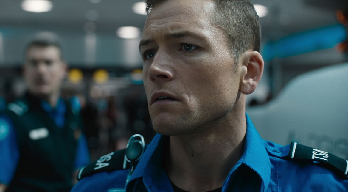 Auf dem Bild aus dem Film Carry-On ist Taron Egerton als Ethan Kopek zu sehen. Er trägt eine blaue Uniform mit TSA-Aufschrift und wirkt ernst und konzentriert. Im unscharfen Hintergrund ist eine weitere Person in ähnlicher Uniform zu erkennen, die die Flughafenatmosphäre unterstreicht. Die Szene vermittelt Spannung und Dramatik.