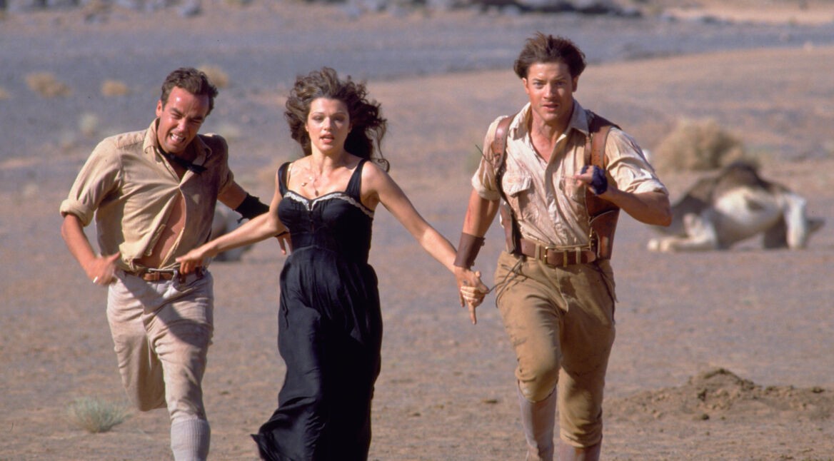 Auf dem Bild aus dem neuen Die Mumie-Film von 1999 sind Brendan Fraser als Rick O'Connell, Rachel Weisz als Evelyn Carnahan und John Hannah als Jonathan Carnahan zu sehen. Die drei rennen durch eine Wüstenlandschaft, sichtbar angespannt und in Eile. Im Hintergrund liegt ein Kamel auf dem Boden. Die Szene vermittelt Action und Spannung.