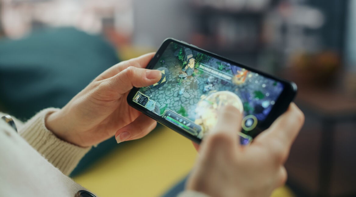 Auf dem Bild ist eine Person zu sehen, die ein Smartphone in beiden Händen hält und ein Strategiespiel spielt. Der Bildschirm zeigt eine farbenfrohe Spielszene mit Figuren und Steinelementen. Die entspannte Umgebung im Hintergrund deutet auf Freizeit hin. Das Bild thematisiert Handyspiele für Erwachsene.