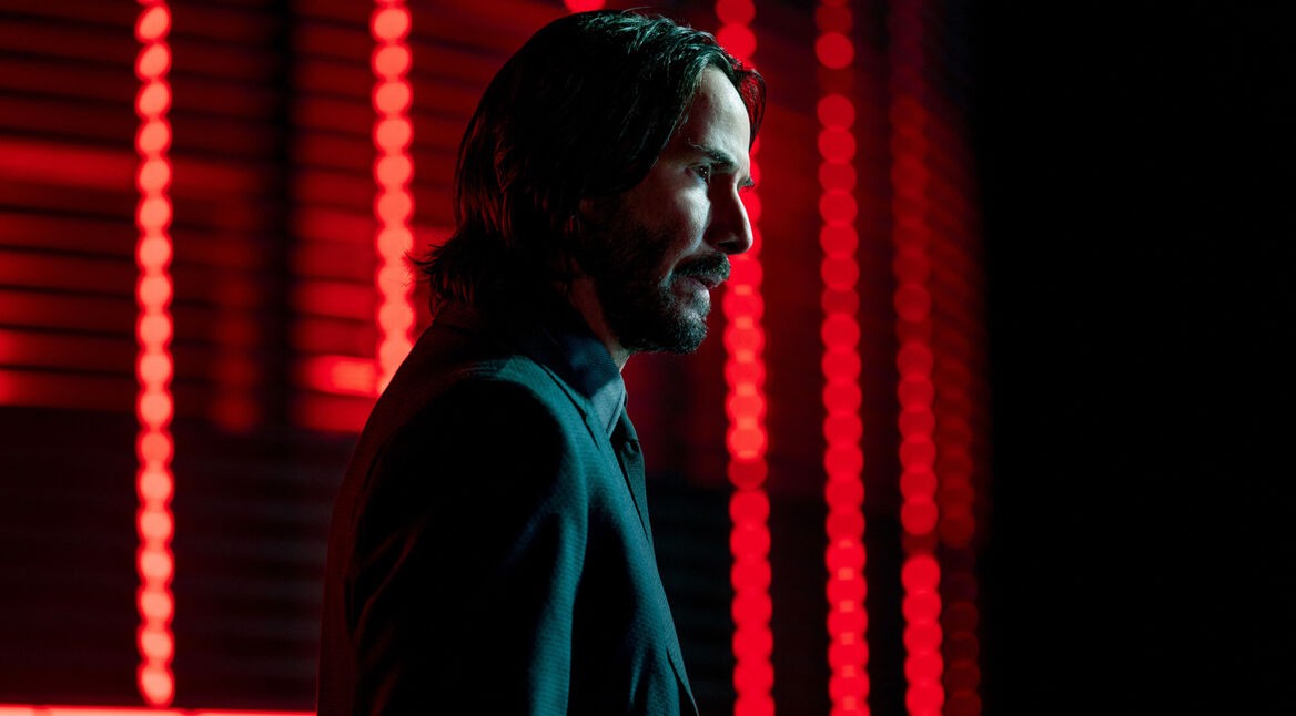 Auf dem Bild in dem Artikel zu den John Wick-Zitaten ist John Wick, gespielt von Keanu Reeves, in einem dunklen Anzug zu sehen. Er steht im Profil vor einem Hintergrund mit leuchtend roten Lichtern. Sein Blick ist ernst und fokussiert, die Szene wirkt stilisiert und atmosphärisch düster, typisch für die Ästhetik der John Wick-Filme.