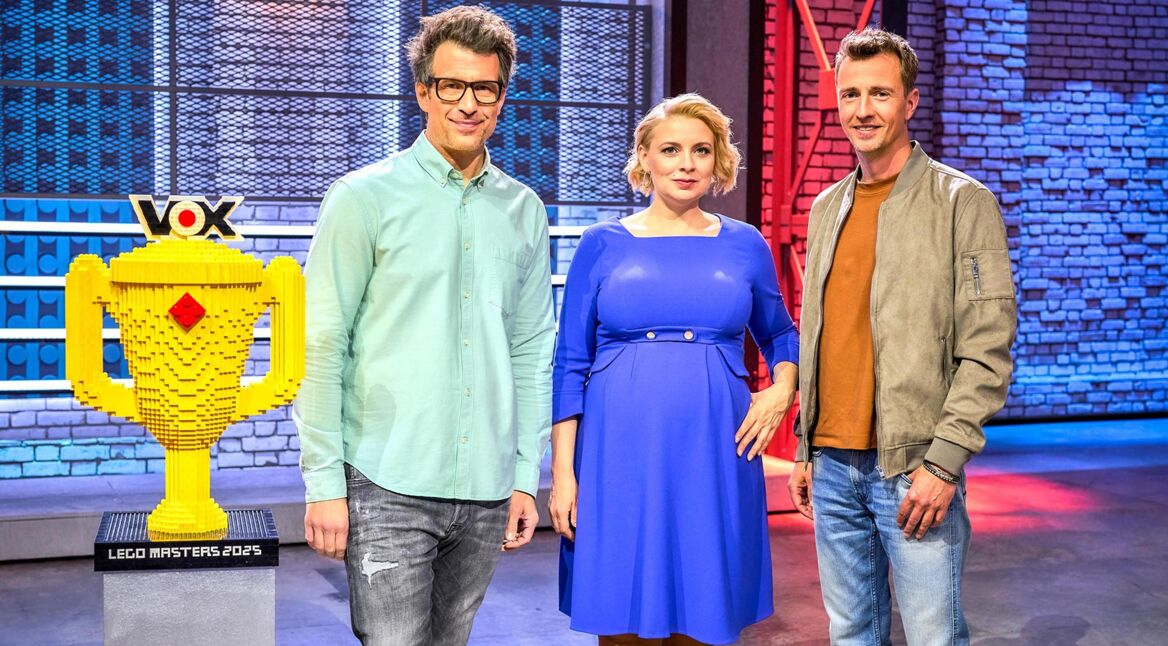 Auf dem Bild zu LEGO Masters 2025 sind der Moderator und die Jury der Show zu sehen. Links steht der Moderator in einem mintgrünen Hemd und mit Brille. In der Mitte befindet sich eine Jurorin in einem blauen Kleid, rechts ein Juror in einer braunen Jacke mit orangefarbenem Shirt. Neben ihnen steht eine große, gelbe LEGO-Trophäe mit dem VOX-Logo. Der Hintergrund zeigt eine moderne Studiokulisse mit blauen und roten Lichteffekten.