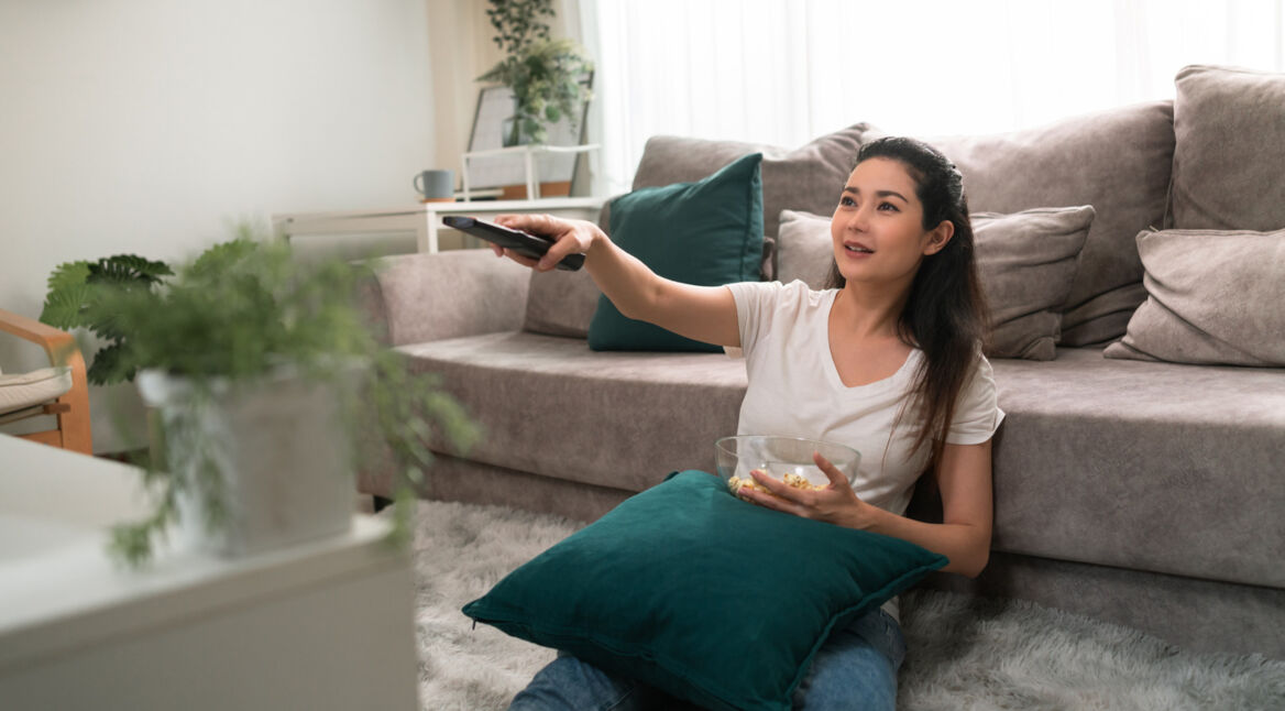 Eine Frau sitzt vor ihrem Sofa auf dem Boden und bedient ihren Fernseher mit der Fernbedienung.