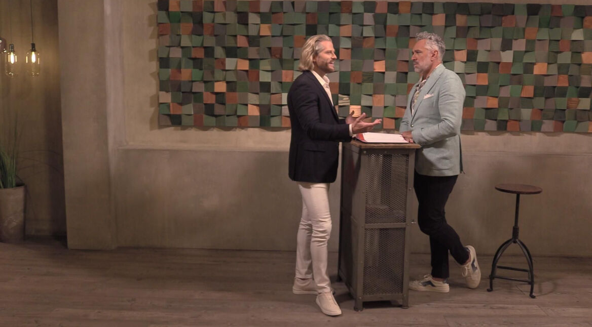 In diesem Bild aus „Promi First Dates“ ist Paul Janke zu sehen, der in einem dunklen Sakko, einem weißen Hemd und weißen Hosen an einem Tresen steht. Er unterhält sich gestikulierend mit einem Mann in einem hellblauen Sakko und weißen Sneakern. Im Hintergrund ist eine Wand mit einer bunten, geometrischen Dekoration zu erkennen, die dem Raum eine moderne Atmosphäre verleiht.