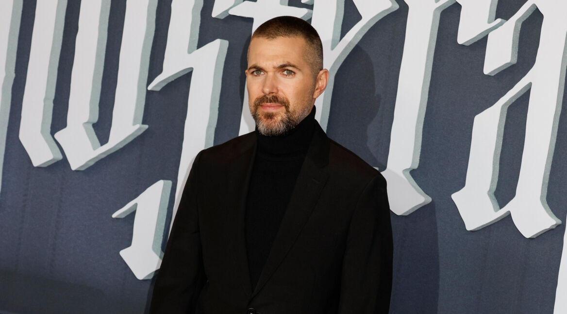 Auf dem Bild ist der Filmemacher Robert Eggers zu sehen, bekannt für seine atmosphärischen Werke. Er trägt einen schwarzen Rollkragenpullover und einen schwarzen Blazer, während er vor einem Hintergrund mit großen, gotischen Buchstaben posiert.