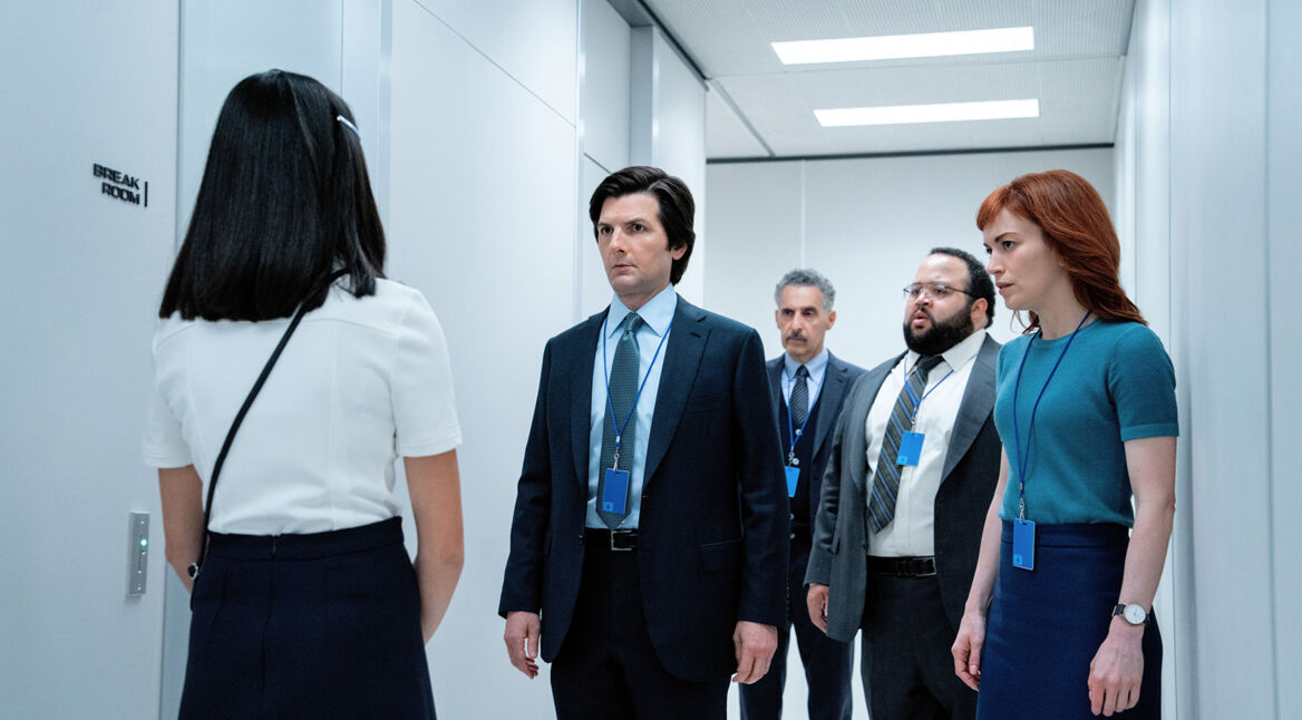 Auf dem Bild aus Severance Staffel 2 stehen von links nach rechts Miss Huang, gespielt von Sarah Bock, Mark Scout, gespielt von Adam Scott, Irving Bailiff, gespielt von John Turturro, Dylan George, gespielt von Zach Cherry, und Helly Riggs, gespielt von Britt Lower, in einem sterilen Büroflur. Alle tragen Bürokleidung mit blauen Namensschildern. Miss Huang steht mit dem Rücken zur Kamera, während die anderen ernsthaft und konzentriert wirken. Die weiße Umgebung und das Wort „Break Room“ an der Wand betonen die kontrollierte und angespannte Stimmung.