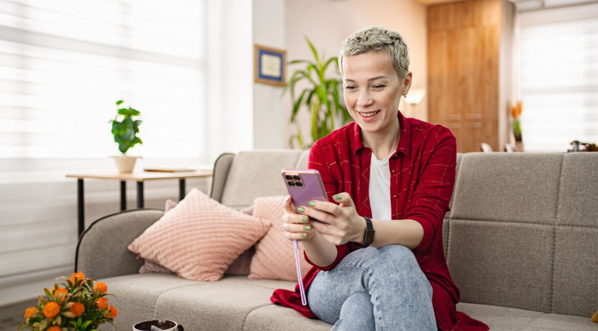 Eine Frau sitzt auf einem Sofa im Wohnzimmer und bedient ein Smartphone.