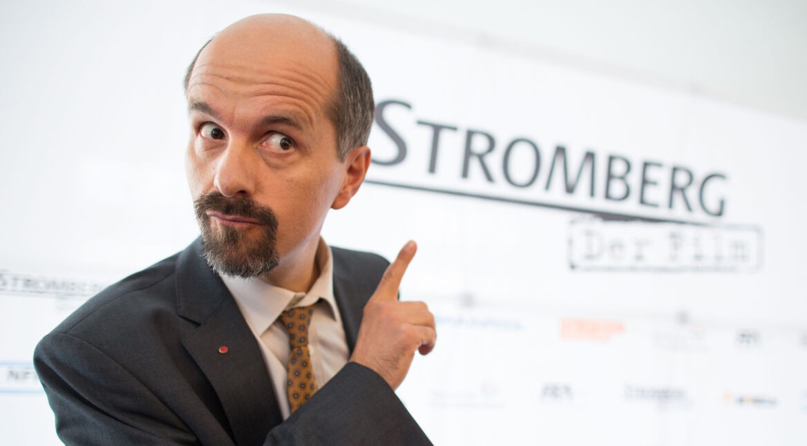 Auf dem Bild zum Stromberg Film 2025 ist Christoph Maria Herbst in seiner Rolle als Bernd Stromberg zu sehen. Er trägt einen dunklen Anzug mit Krawatte und zeigt mit erhobenem Zeigefinger auf den Schriftzug "Stromberg – Der Film" im Hintergrund. Sein Gesichtsausdruck ist verschmitzt und charakteristisch für die Figur.