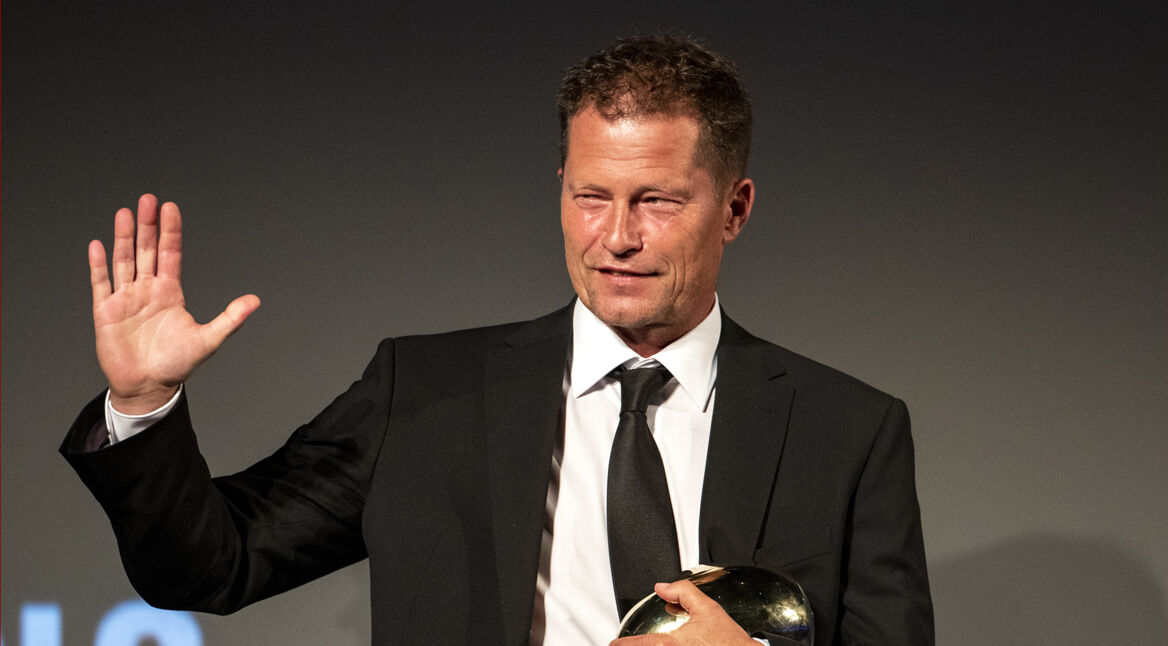 Auf dem Bild zum Film The Neglected ist Til Schweiger zu sehen, der einen schwarzen Anzug mit weißem Hemd und schwarzer Krawatte trägt. Er hebt die rechte Hand lächelnd zum Gruß und hält mit der anderen Hand eine glänzende goldene Trophäe. Der Hintergrund ist dunkel, was den Fokus auf Schweiger legt.