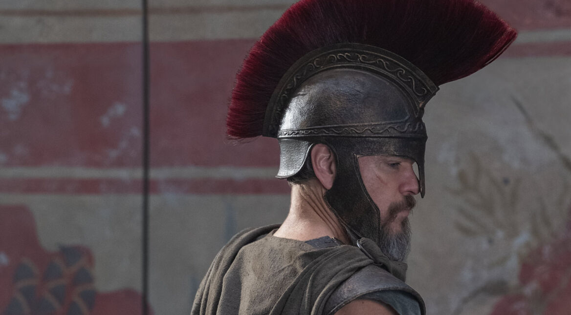 Das Bild aus The Odyssey zeigt Matt Damon in der Rolle des Odysseus. Er trägt eine antike Rüstung mit einem markanten bronzenen Helm, der mit einem roten Kamm verziert ist. Sein Gesichtsausdruck ist ernst und fokussiert, während er zur Seite blickt. Der Hintergrund zeigt eine Wand mit verblassten Fresken, die an antike griechische Kunst erinnern. Die Szene vermittelt eine epische und historische Atmosphäre, passend zur legendären Geschichte von Odysseus.