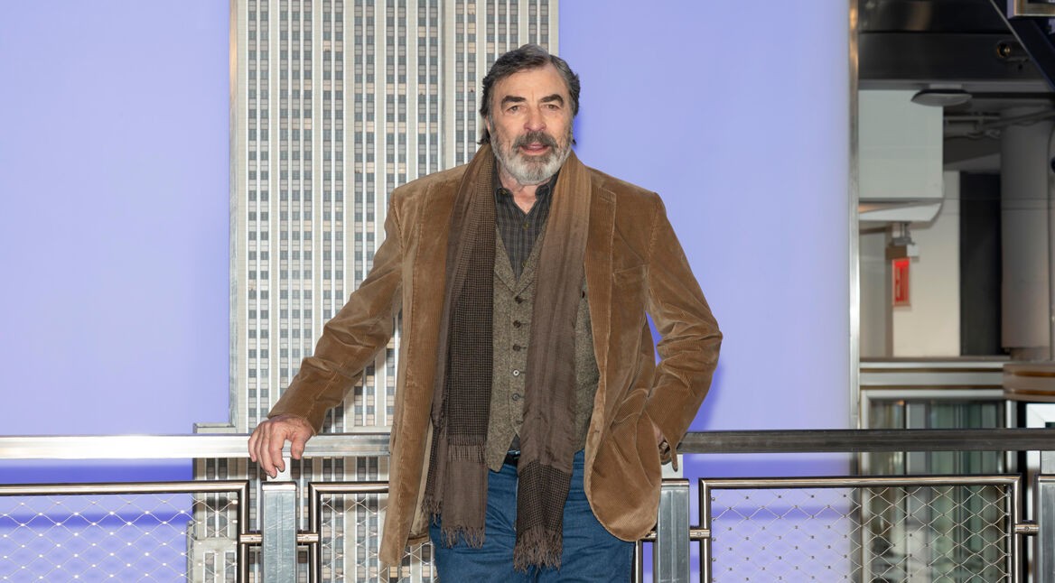 Auf dem Bild ist Tom Selleck zu sehen, wie er lässig an einem Geländer lehnt. Er trägt eine braune Cordjacke, ein gemustertes Hemd und einen Schal in Erdtönen. Sein Blick ist freundlich und selbstbewusst, im Hintergrund ragt ein Teil eines Gebäudes in die Höhe vor einem bläulichen Himmel. Das Setting wirkt urban und stilvoll.