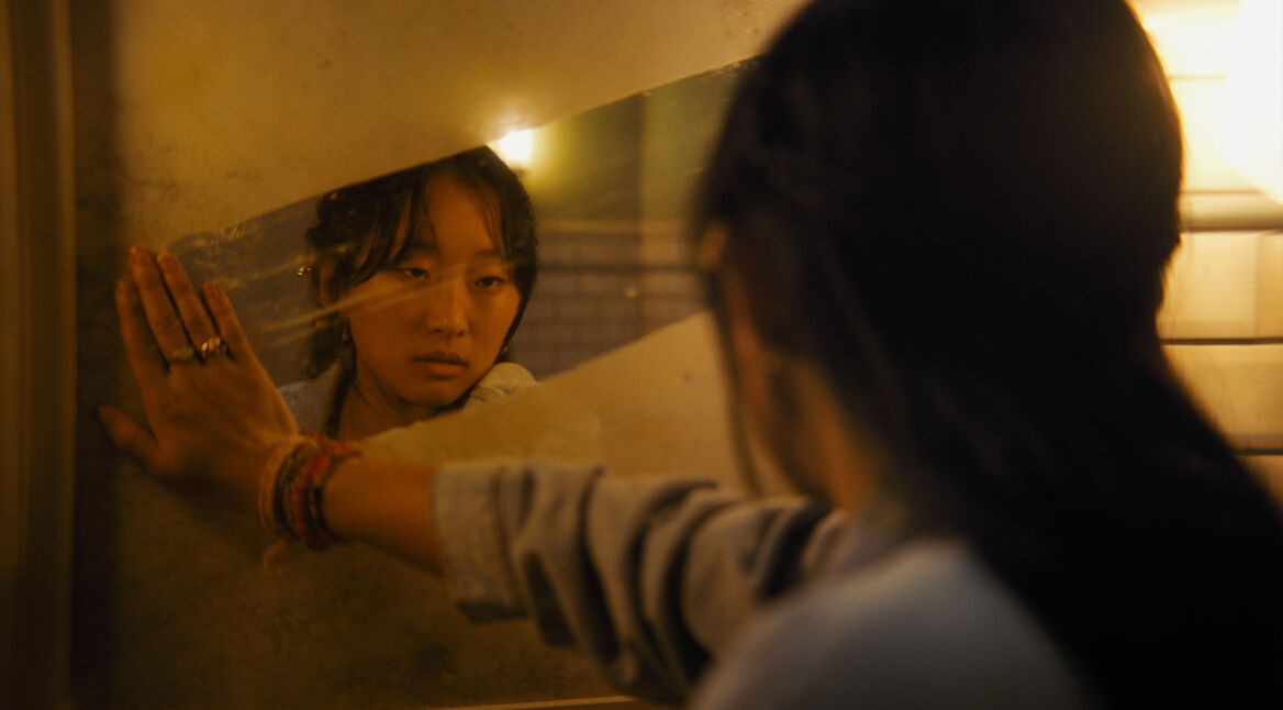 Im Bild zum Until Dawn-Film ist Megan, gespielt von Ji-young Joo, zu sehen. Sie blickt ernst in einen schmutzigen Spiegel, während ihre Hand gegen die spiegelnde Oberfläche drückt. Die Szene ist in warmes, gedämpftes Licht getaucht, was eine nachdenkliche und emotionale Stimmung erzeugt.