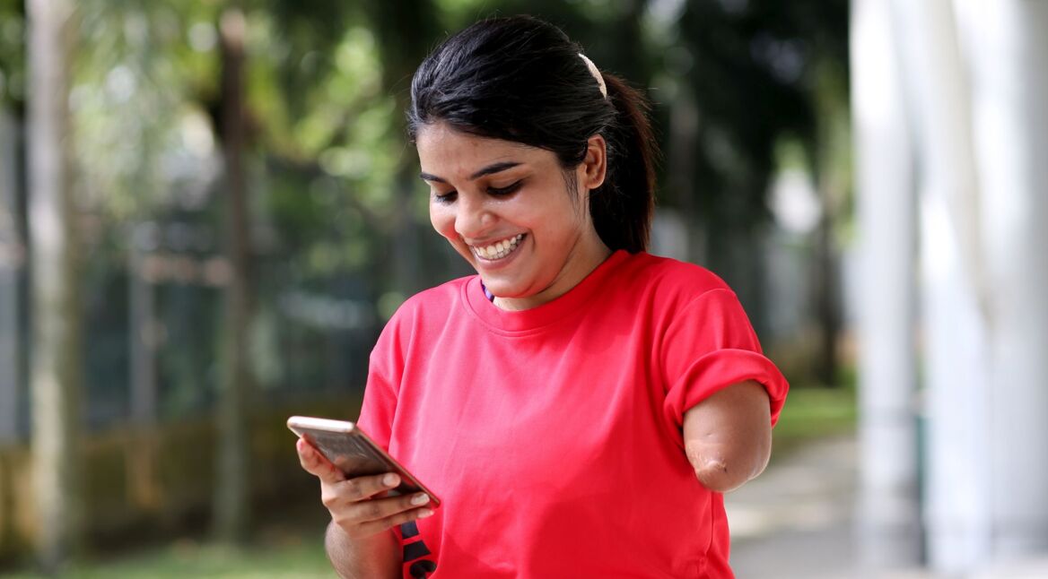 Junge Frau schaut lächelnd auf ein Smartphone, das sie in der rechten Hand hält.