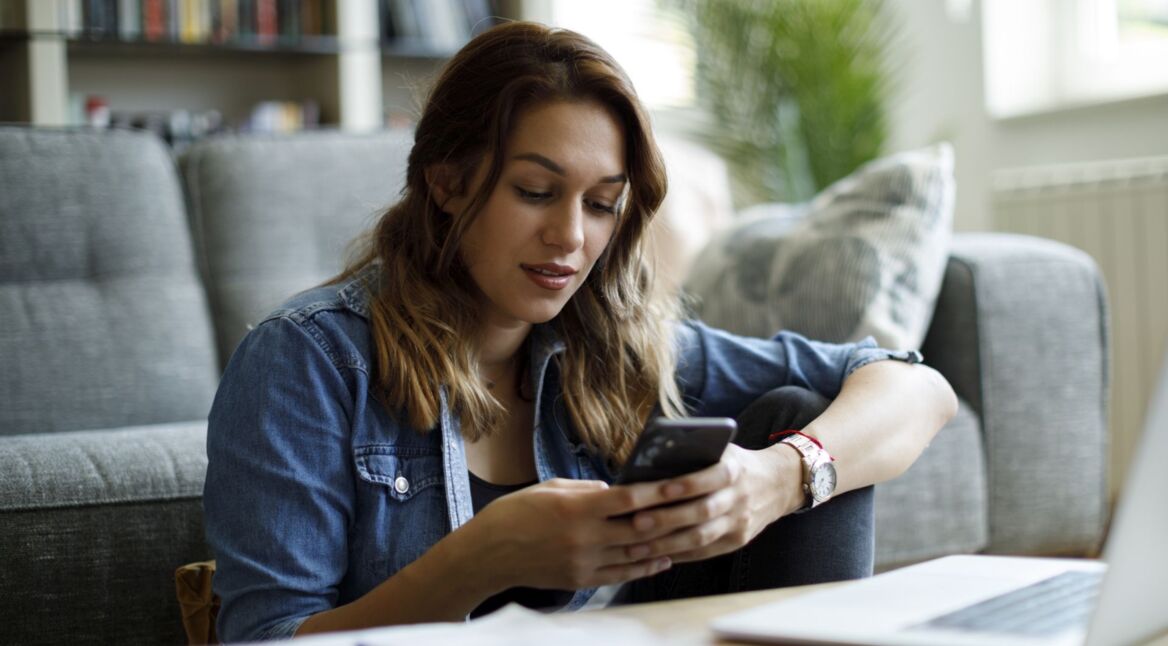 Junge Frau sitzt auf dem Boden vor ihrem Sofa und schaut lächelnd auf ihr Android-Handy.