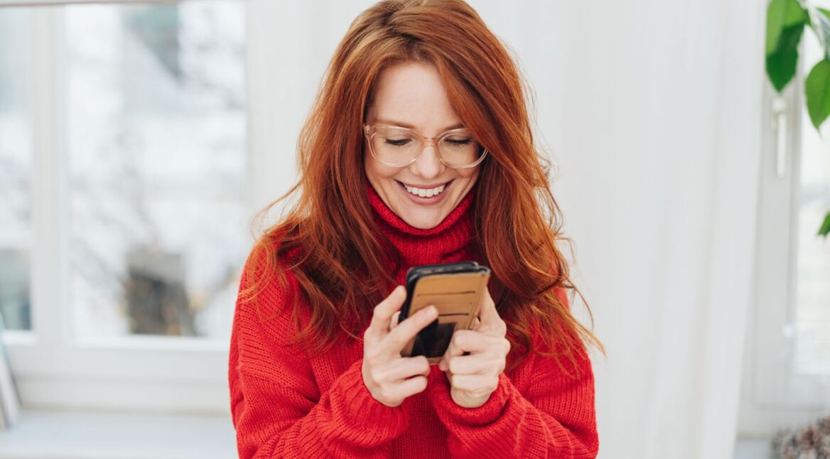 Rothaarige junge Frau in rotem Pullover schaut auf Android-Handy, das sie in den Händen hält.