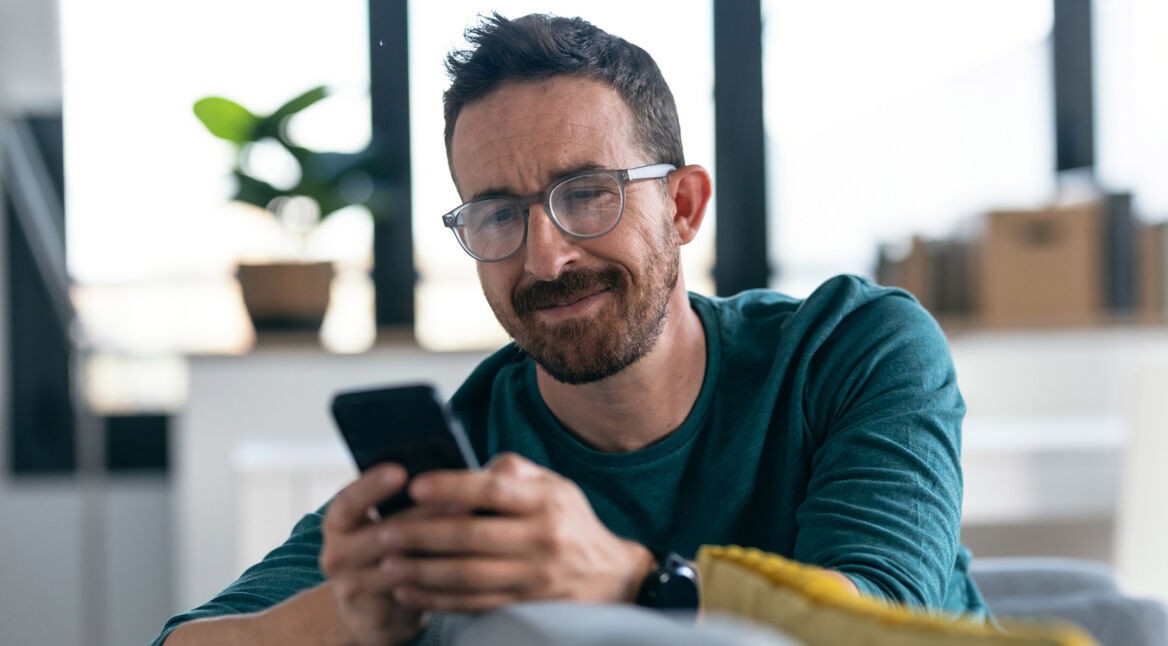 Ein Mann sitzt auf einem Sofa und schaut auf ein Smartphone, das er in seinen Händen hält