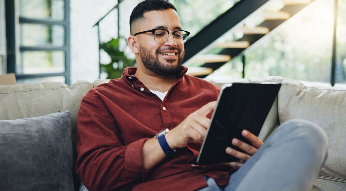 Junger Mann sitzt auf der Couch und arbeitet an seinem Tablet.