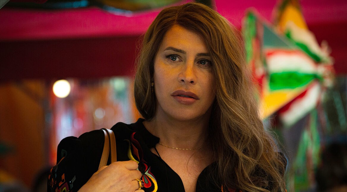 Auf dem Bild aus Emilia Pérez ist Karla Sofía Gascón in der Rolle von Emilia Pérez zu sehen. Sie trägt eine schwarze Bluse mit bunten Stickereien und hält eine Tasche über der Schulter. Ihr Gesichtsausdruck ist ernst und fokussiert, während sie in eine farbenfrohe, belebte Umgebung blickt. Die Szene vermittelt eine angespannte, nachdenkliche Stimmung. Emilia Pérez streamen ermöglicht es, diesen Moment hautnah mitzuerleben.