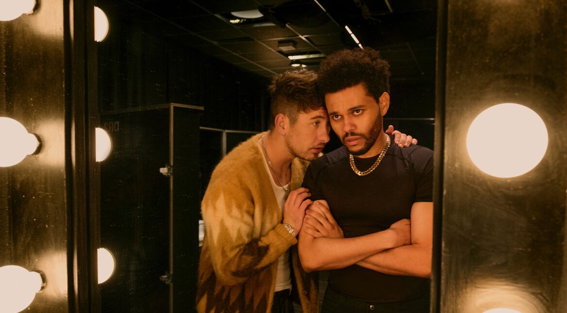 Auf dem Bild aus Hurry Up Tomorrow Film sind Barry Keoghan und Abel Tesfaye (The Weeknd) in einer intensiven Szene zu sehen. Barry Keoghan, in einer gemusterten Jacke, lehnt sich nah an Tesfaye heran und flüstert ihm etwas zu, während er eine ernste Miene zeigt. Abel Tesfaye steht mit verschränkten Armen vor einem Spiegel mit hellen Glühbirnen und wirkt angespannt. Die dunkle Garderobenraum-Atmosphäre verstärkt die dramatische Stimmung.