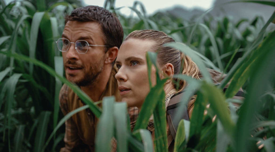 Auf dem Bild siehst Du eine Szene aus Jurassic World: Die Wiedergeburt. Links ist Jonathan Bailey als Paläontologe Dr. Henry Loomis zu sehen, rechts Scarlett Johansson als die verdeckte Einsatzspezialistin Zora Bennett. Beide ducken sich in dichtem, grünen Gras und blicken konzentriert in die Ferne, als würden sie eine Bedrohung beobachten.
