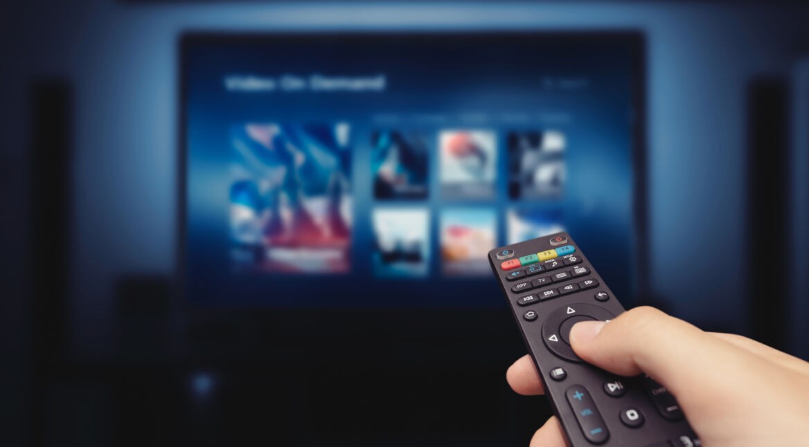 Auf dem Bild in dem Artikel zu den Prime Video-Kosten hält eine Person eine schwarze Fernbedienung in der Hand und navigiert durch das Menü eines Streaming-Dienstes auf einem großen Bildschirm. Die Oberfläche zeigt verschiedene Filme und Serien zur Auswahl. Die Szene ist in dunklen Blau- und Schwarztönen gehalten, was eine gemütliche Heimkino-Atmosphäre erzeugt.