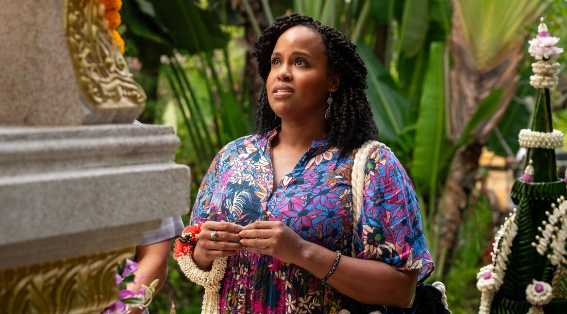 Auf dem Bild aus The White Lotus Staffel 3 ist Natasha Rothwell in Episode 1 als Belinda Lindsey zu sehen. Sie trägt ein farbenfrohes, blumiges Kleid und blickt nachdenklich auf eine verzierte Steinstruktur. In ihren Händen hält sie Blumenschmuck. Der Hintergrund ist von tropischen Pflanzen und traditionellen Dekorationen geprägt, was die exotische Atmosphäre unterstreicht. Der The White Lotus Staffel 3 Cast umfasst unter anderem ihre Rolle.