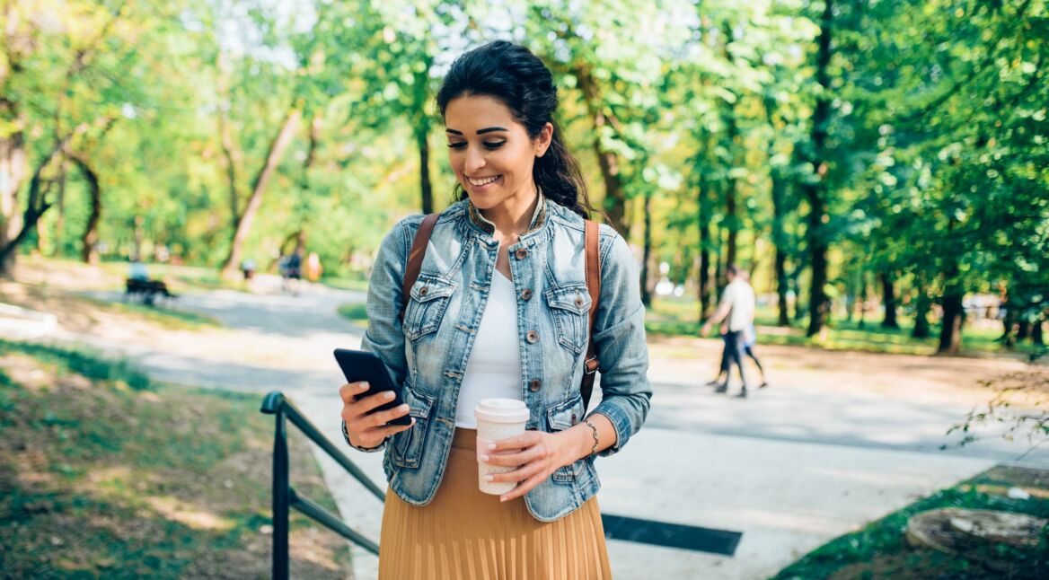 Frau steht im Park mit ihrem Smartphone und einem Kaffee in der Hand.
