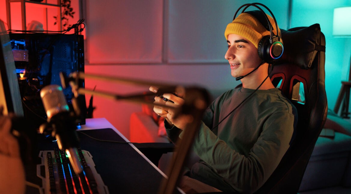 Ein junger Mann sitzt auf einem PC-Stuhl, hält einen Controller in der Hand und spielt am Computer.