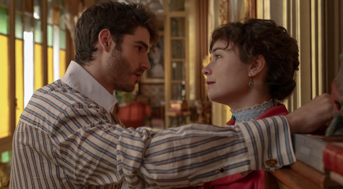 In dieser Szene aus Handbuch einer Anstandsdame Staffel 2 stehen sich ein junger Mann und eine junge Frau in einem prunkvoll eingerichteten Raum gegenüber. Der Mann lehnt mit einem Arm an der Wand und blickt intensiv in die Augen der Frau, die ruhig und ernst erwidert. Die Stimmung wirkt romantisch und gespannt zugleich.