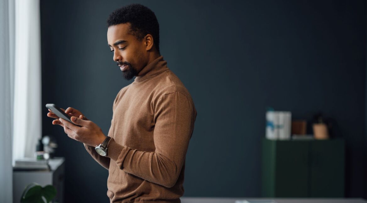 Junger Mann steht in einem Raum und schaut auf sein Smartphone.
