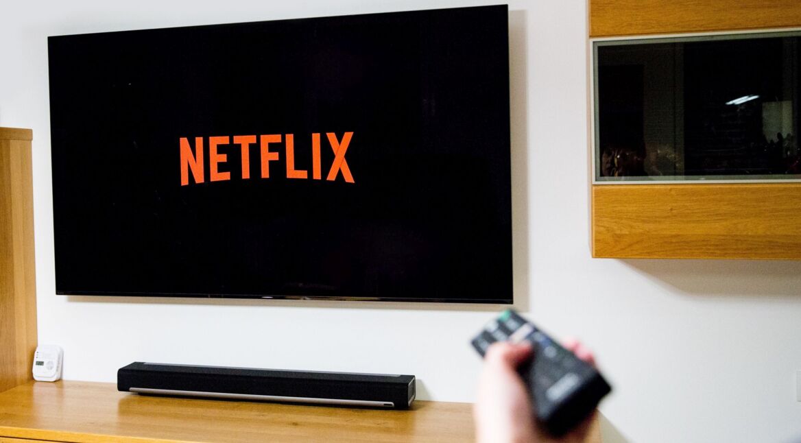 Eine Person sitzt im Wohnzimmer und schaltet mit der Fernbedienung Netflix auf dem TV ein.