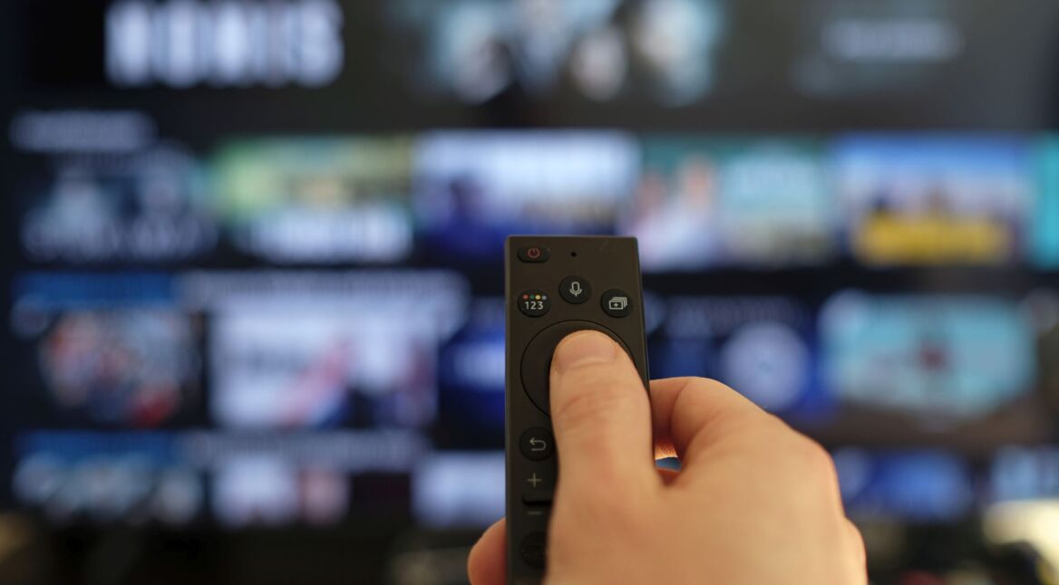 Hand hält eine Fernbedienung vor einen Fernseher