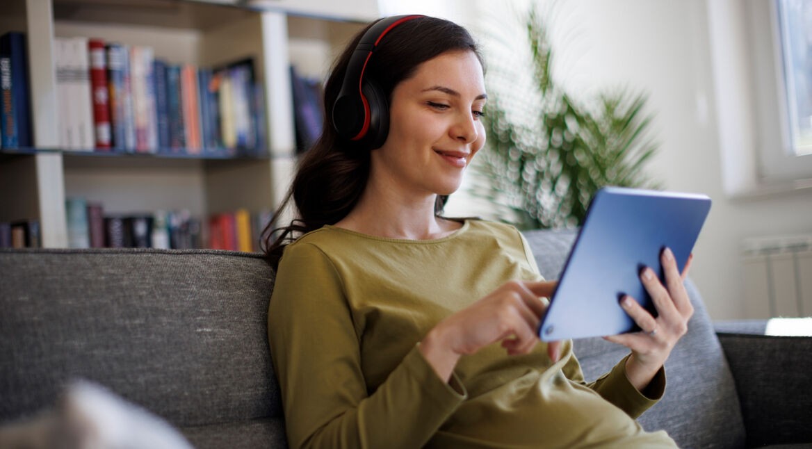 Auf dem Bild ist eine junge Frau mit dunklen Haaren zu sehen, die entspannt auf einem grauen Sofa sitzt. Sie trägt schwarze Kopfhörer mit roten Akzenten und ein olivgrünes Oberteil. In ihren Händen hält sie ein blaues Tablet und tippt darauf, während sie zufrieden lächelt. Im Hintergrund steht ein Bücherregal mit bunten Büchern. Das Bild vermittelt eine gemütliche Atmosphäre, passend zur Nutzung von Arthaus Plus für Streaming.