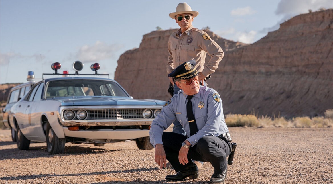 In Dark Winds Staffel 3 sind Lt. Joe Leaphorn (Zahn McClarnon) und Gordo Sena (A. Martinez) in einer wüstenartigen Landschaft zu sehen. Leaphorn steht mit Sonnenbrille und Hut neben einem alten Polizeiwagen, während Sena in blauer Uniform und mit ernster Miene in der Hocke verharrt.