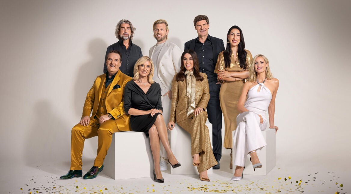 Auf dem Bild zur Sendung Die Höhle der Löwen 2025 sind die Juror:innen in eleganter Kleidung vor einem neutralen Hintergrund zu sehen. Sie tragen goldene, schwarze und weiße Outfits, passend zum luxuriösen Stil der Show. Die Gruppe sitzt und steht in einer harmonischen Formation, umgeben von goldenen Konfetti-Elementen auf dem Boden, was eine festliche Stimmung vermittelt.
