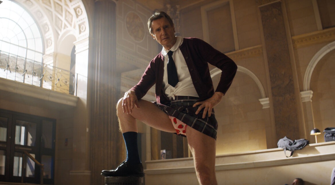 In dieser Szene aus Die nackte Kanone 4 von 2025 steht Liam Neeson als Frank in einem prachtvollen, klassizistischen Gebäude mit einem Bein auf einem Podest. Er trägt eine Schuluniform mit offenem Hemd, Krawatte, kariertem Rock und sichtbar roten Herzchen-Boxershorts, was der Szene eine absurde und humorvolle Stimmung verleiht.