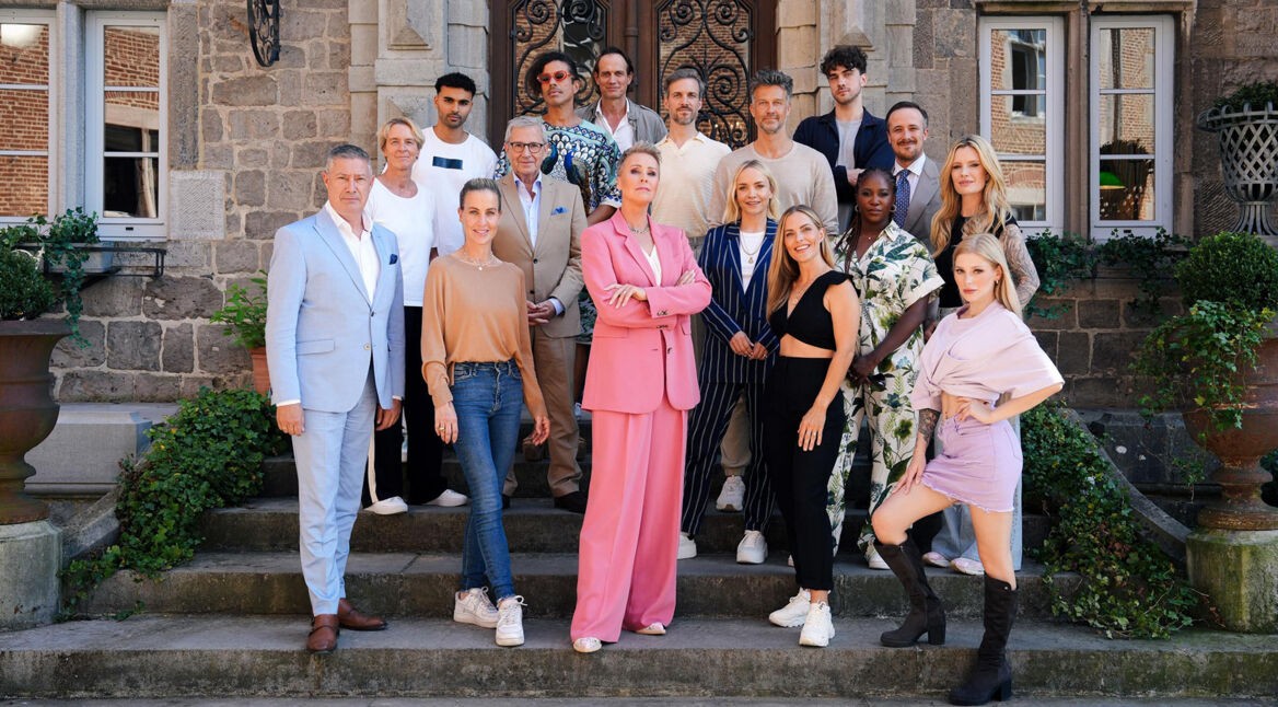 Auf dem offiziellen Gruppenfoto zu Die Verräter Staffel 3 stehen 16 Teilnehmer:innen und die Moderatorin auf einer Steintreppe vor einem historischen Gebäude. Im Mittelpunkt steht eine Frau im auffälligen pinken Hosenanzug mit verschränkten Armen. Die Gruppe wirkt selbstbewusst, gespannt und bereit für das Spiel.