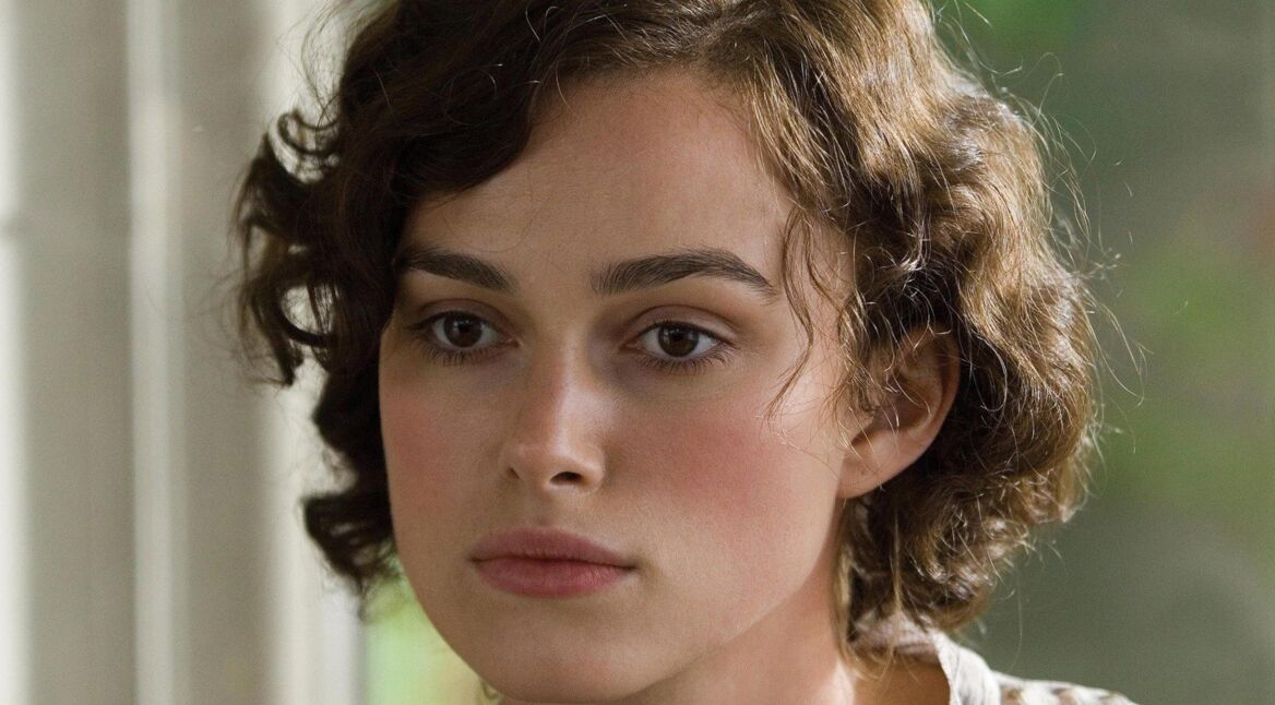 Auf diesem Bild aus dem Film Abbitte ist Keira Knightley in der Rolle der Cecilia Tallis zu sehen. Sie blickt nachdenklich aus dem Fenster, ihr Gesicht wirkt ruhig, aber innerlich bewegt. Ihr braunes, leicht gelocktes Haar ist kurz geschnitten, und die Szene vermittelt eine melancholische, ernste Stimmung. Ideal für eine Übersicht zu Filme mit Keira Knightley.