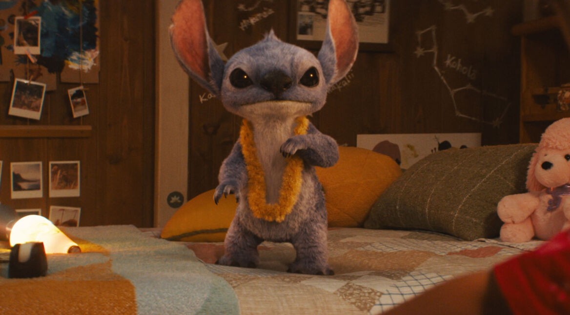Auf dem Bild aus der Lilo und Stitch Live-Action-Adaption 2025 ist das blaue Alien Stitch in einer realistischen CGI-Version zu sehen. Er steht auf einem Bett, trägt eine gelbe Blumenlei und blickt direkt in die Kamera.