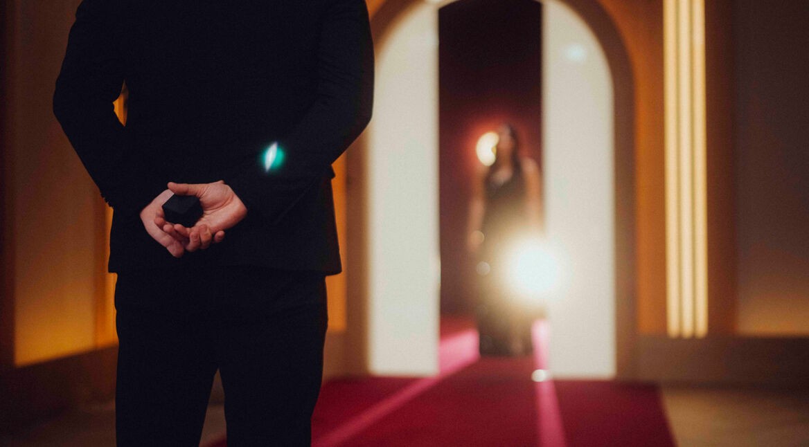 Auf dem Bild in dem Artikel zu Love Is Blind: Germany Staffel 2 steht ein Mann im schwarzen Anzug mit dem Rücken zur Kamera auf einem roten Teppich und hält eine kleine schwarze Schachtel hinter dem Rücken. Im Hintergrund erscheint eine Frau im Abendkleid.