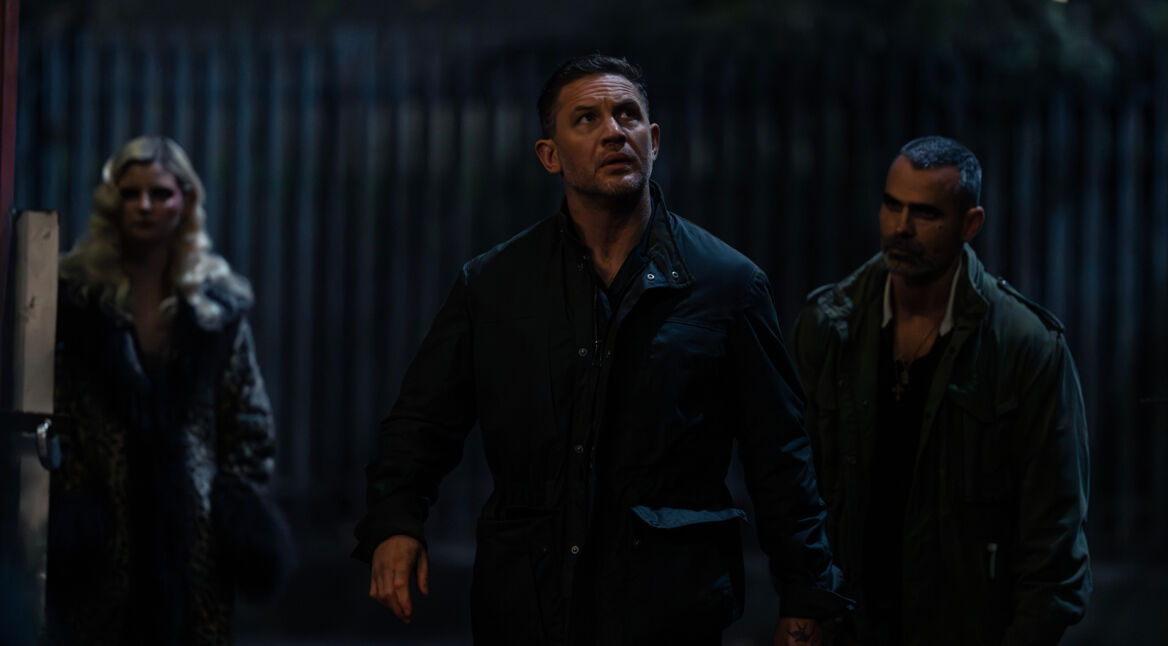 Auf dem Bild zur Mobland Serie ist Tom Hardy als Harry Da Souza zu sehen. Er steht in einer dunklen Gasse und blickt angespannt nach oben, als ob er eine Bedrohung wahrnimmt. Er trägt eine dunkle Jacke, und sein Gesichtsausdruck wirkt fokussiert. Hinter ihm stehen zwei weitere Personen – eine Frau mit blondem Haar in einem auffälligen Mantel und ein Mann mit kurz rasiertem Haar und einem ernsten Blick. Die Szene ist düster beleuchtet und vermittelt eine gespannte Atmosphäre.