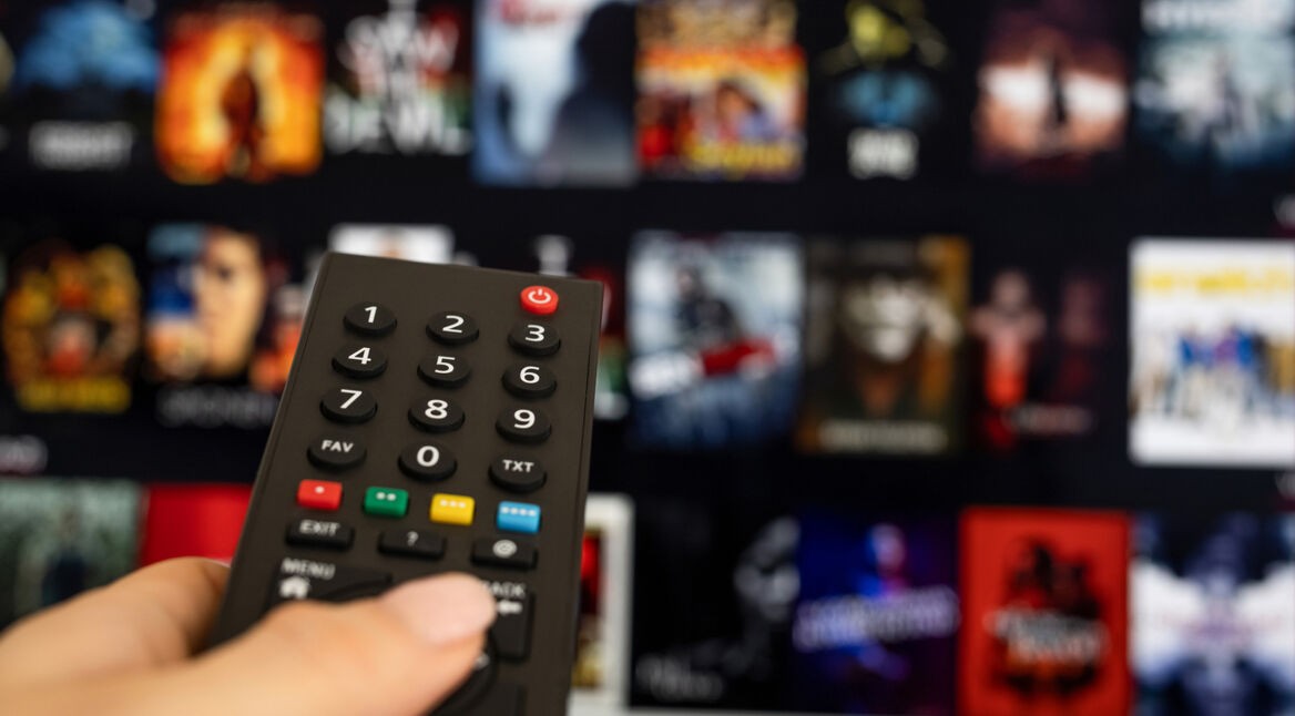 Auf dem Bild ist eine Hand zu sehen, die eine schwarze Fernbedienung hält und auf einen Fernseher zeigt. Der Bildschirm im Hintergrund zeigt die Benutzeroberfläche eines Streaming-Dienstes mit verschiedenen Film- und Serien-Covern, die leicht unscharf sind. Das Bild vermittelt eine typische Streaming-Situation und passt zum Thema Heimkino. Paramount+ könnte als einer der verfügbaren Dienste dargestellt sein.